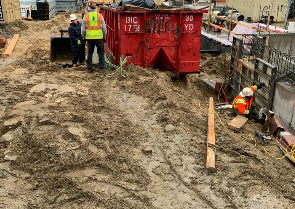 ### Key Elements**Main Subjects** - Construction workers in safety gear - Large red dumpster**Objects** - Construction equipment (e.g., shovel) - Dirt and debris at the construction site**Notable Features** - Safety vests worn by workers - Excavation area with visible trench**Text/Symbols** - Markings on the dumpster (e.g., "30 YD")### Overall Mood/Theme The image conveys a busy construction site atmosphere, highlighting the labor-intensive nature of building projects. The presence of workers and heavy machinery suggests active work, while the safety gear reflects a focus on safety amidst the chaotic environment. The earthy tones of the dirt and debris contribute to a gritty, industrious feel.